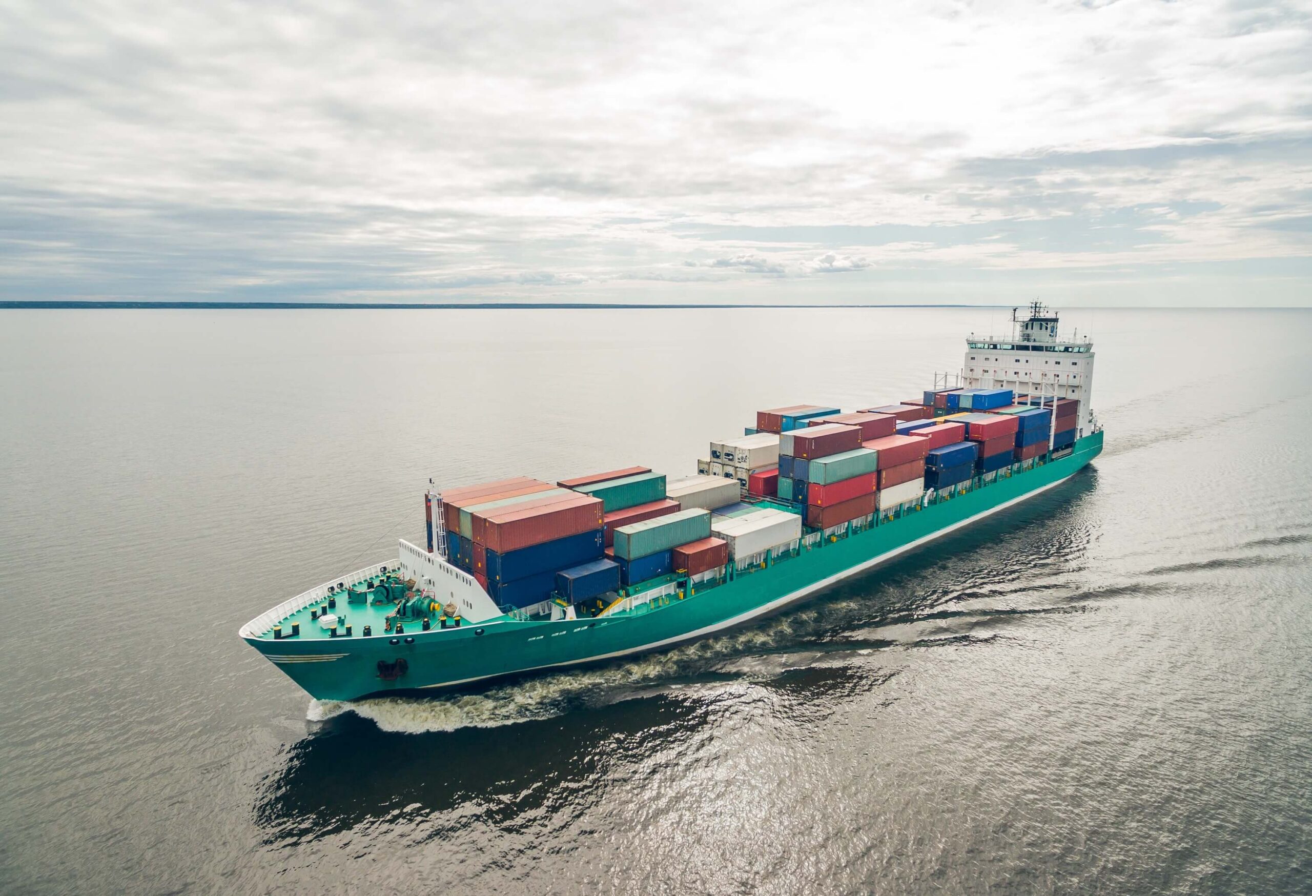 A large cargo ship with vessel clearance, carrying colorful shipping containers, sails across calm waters under a cloudy sky, leaving a wake behind it (CBP rulings and Jones Act).