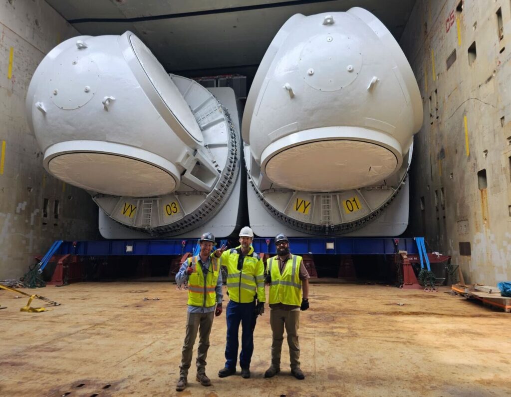 Three workers in safety gear stand in front of two large, white industrial capsules or modules inside a spacious facility or warehouse. The capsules are labeled "VY 03" and "VY 01.