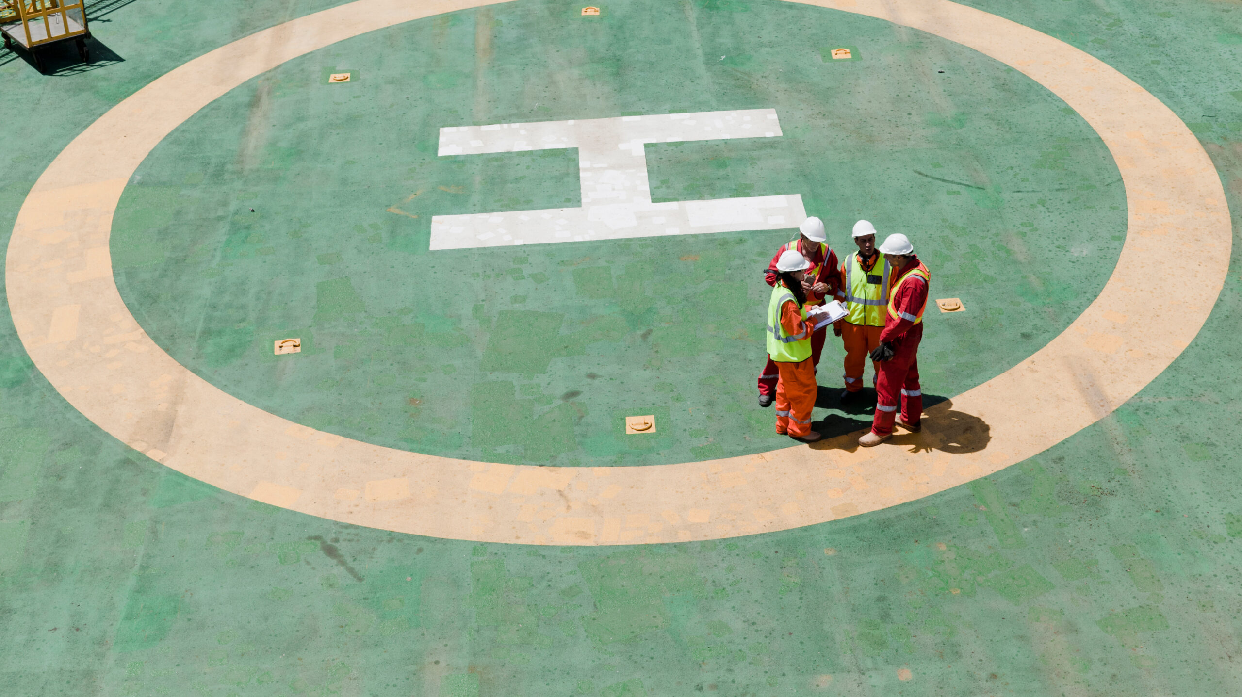Four construction workers wearing safety gear and helmets stand in the center of a large green helipad, discussing documents. The white "H" marking for helicopters is visible on the ground.