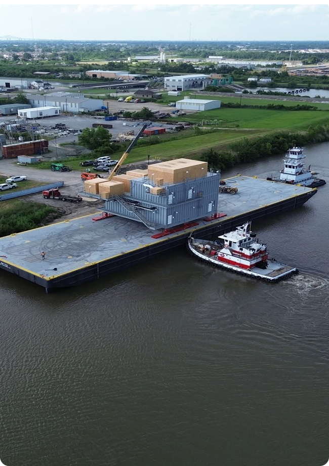 An aerial view of a tugboat guiding a large barge carrying industrial equipment and wooden crates on a river, with a crane and buildings visible on the grassy riverbank.