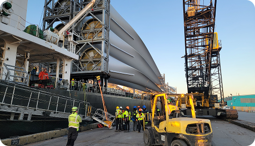 Workers wearing safety gear stand near heavy machinery and a large ship, which is being loaded with several large wind turbine blades using cranes at an industrial dock.