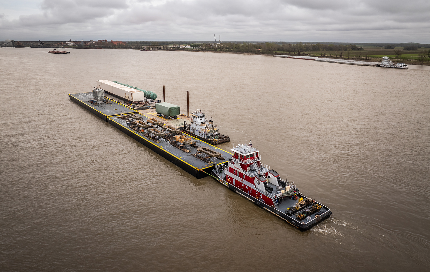 A red and white tugboat pushes a large barge carrying industrial equipment and containers along a wide river, with overcast skies and distant shoreline visible in the background.