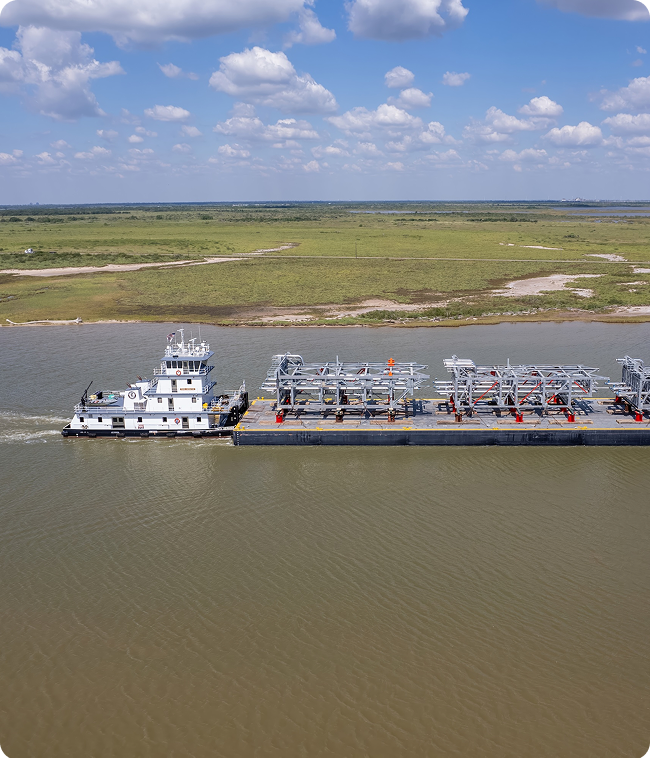 A tugboat pushes a large barge carrying metal structures along a wide river, with grassy shoreline and open sky in the background.