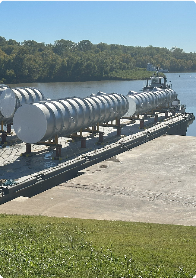 A barge loaded with large cylindrical metal tanks is docked at a ramp by a river, surrounded by green grass and trees under a clear blue sky (barge transportation).