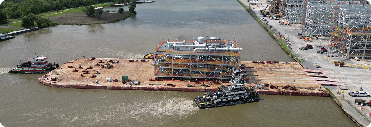 A large industrial module is being transported on a flat barge along a river, guided by tugboats. The barge is near a construction site and surrounded by water and greenery.