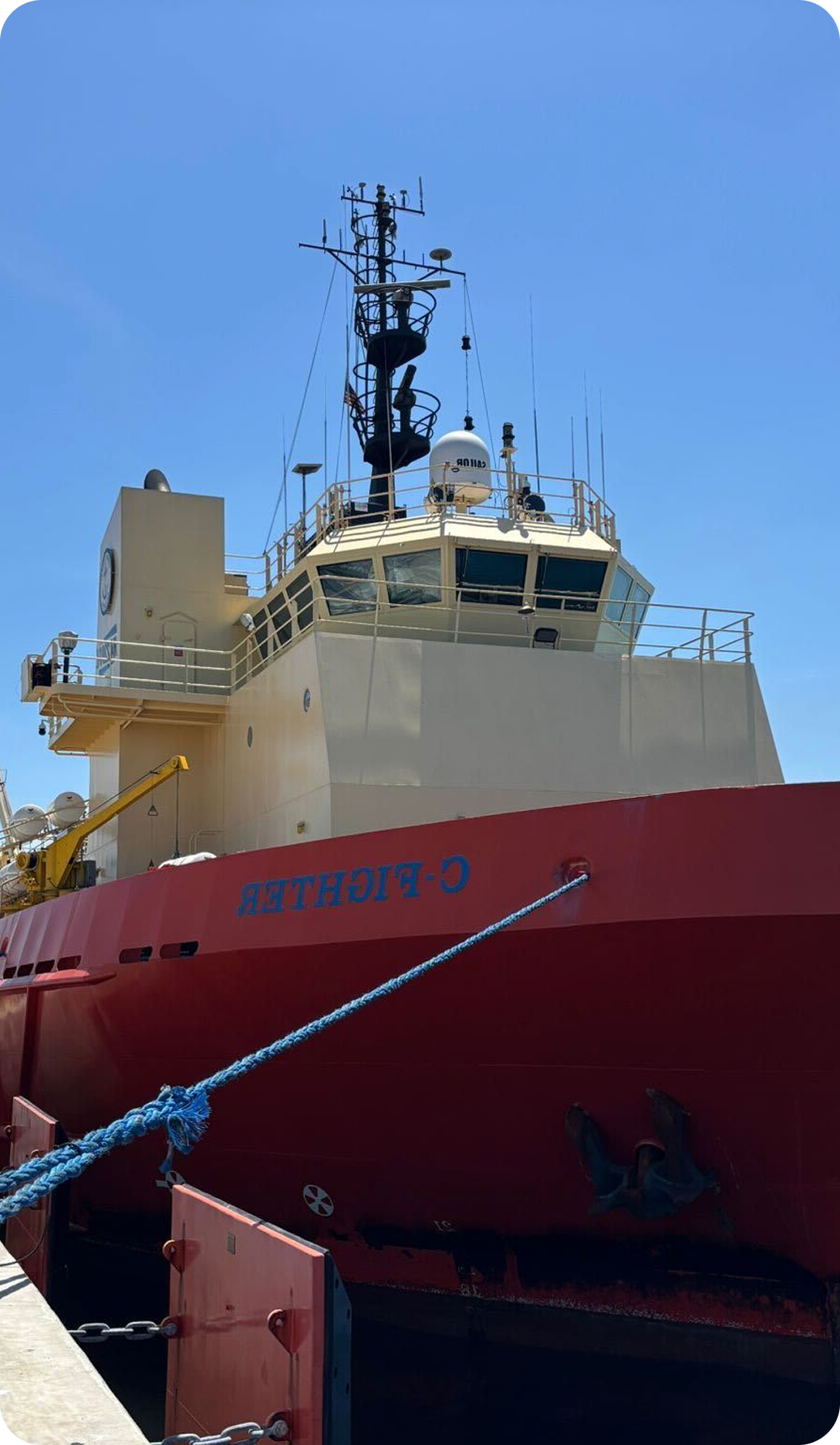 A large red and yellow ship named "O-FIGHTER" is docked at a pier, secured with blue ropes, under a clear blue sky. The ship's anchor and communication equipment are visible.