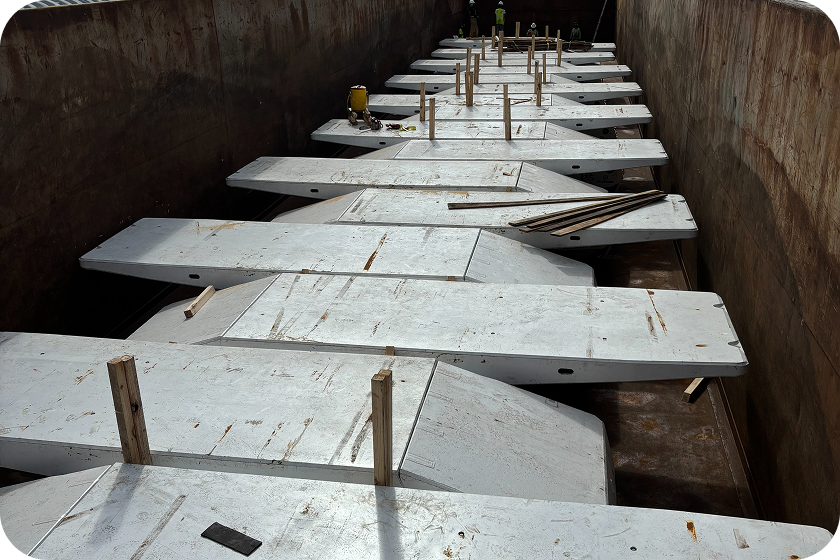 Several large, flat white metal panels with wooden supports are stacked in a row inside a rusted metal container or cargo hold, with construction tools visible on top of the panels.