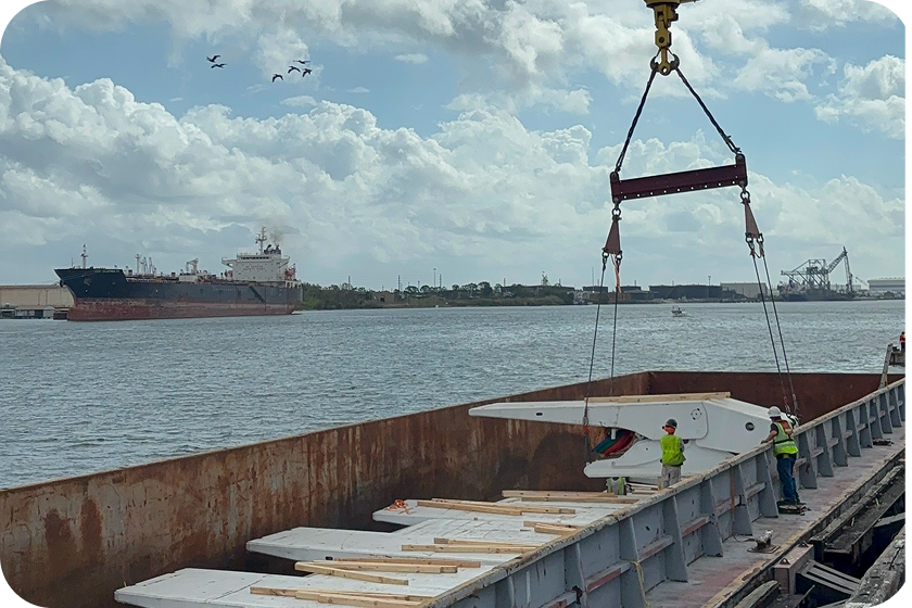 Two workers guide a large white blade being lowered by a crane into a cargo ship on a river. Other blades rest in the hold, with a large ship and birds visible in the background under a partly cloudy sky.