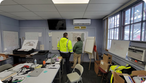 Two people in safety jackets stand by a whiteboard covered with diagrams in a cluttered office with large windows, computers, and various office supplies scattered around.