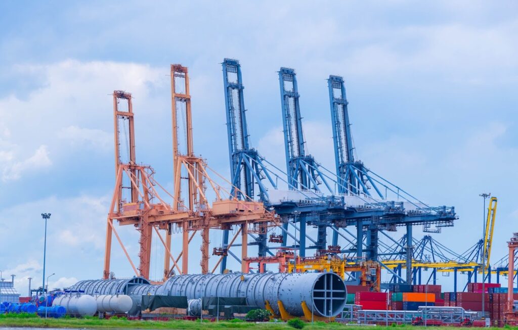 Large shipping cranes stand at a busy port, with shipping containers and cylindrical industrial equipment in the foreground under a cloudy sky—a reminder of the importance of knowing how to avoid demurrage and detention charges.