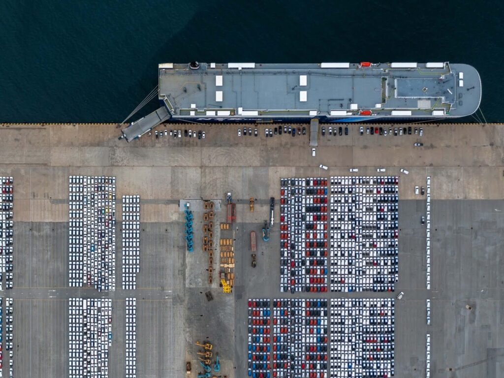 Aerial view of a large cargo ship docked at a port, with rows of parked cars and industrial vehicles—an organized scene that highlights the importance of how to avoid demurrage and detention charges.