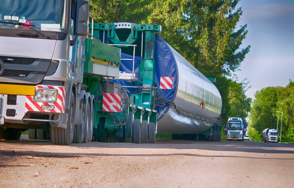 A large truck transports an oversized load—a long cylindrical wind turbine blade—along a purposed route on a rural road, accompanied by support vehicles and surrounded by trees and greenery.