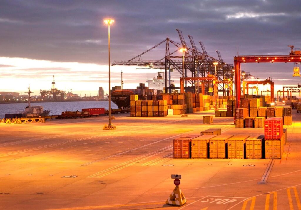 A brightly lit shipping port at sunset with stacked cargo containers, cranes, and industrial equipment—showcasing efficient operations that help shippers learn how to avoid demurrage and detention charges. The sky is partly cloudy, with water in the background.