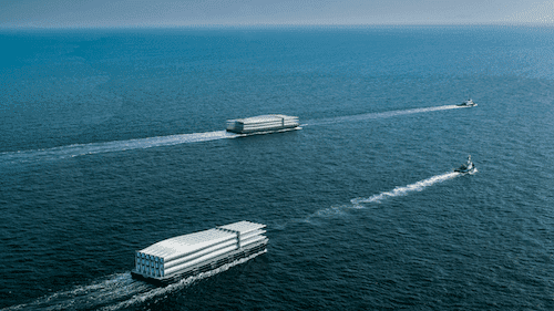 Two large, rectangular, white floating structures are being towed by small boats across a calm, open sea under a clear sky.