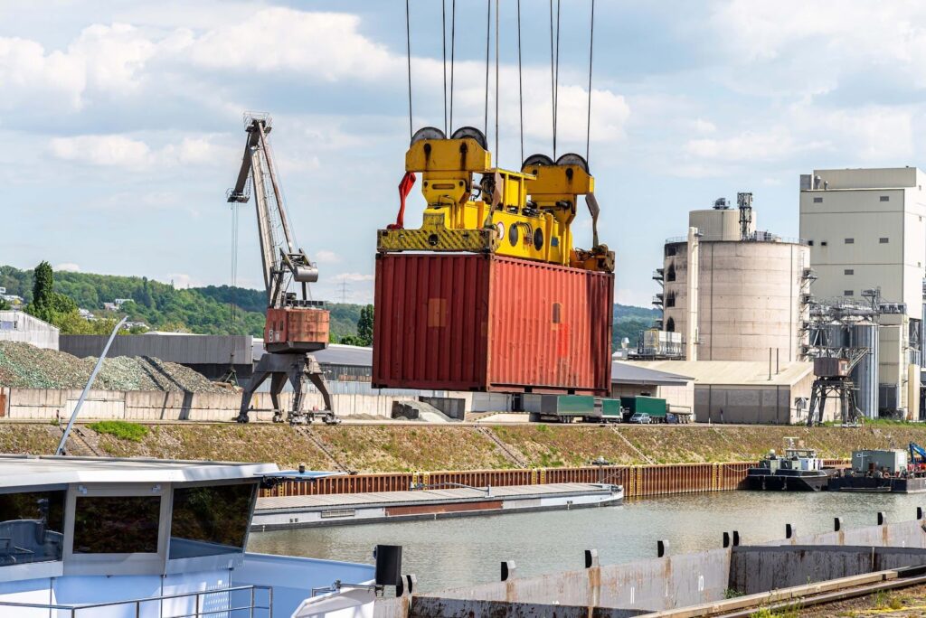 A red shipping container is being lifted by a yellow crane at an industrial port—an essential scene for anyone learning how to avoid demurrage and detention charges, with warehouses, cranes, and a canal visible beneath a partly cloudy sky.