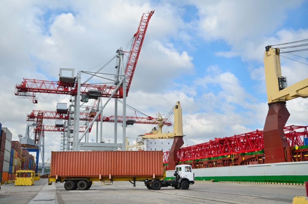 A white truck carries a large shipping container at a busy port surrounded by cranes and stacked containers, illustrating the importance of knowing how to avoid demurrage and detention charges under a partly cloudy sky.