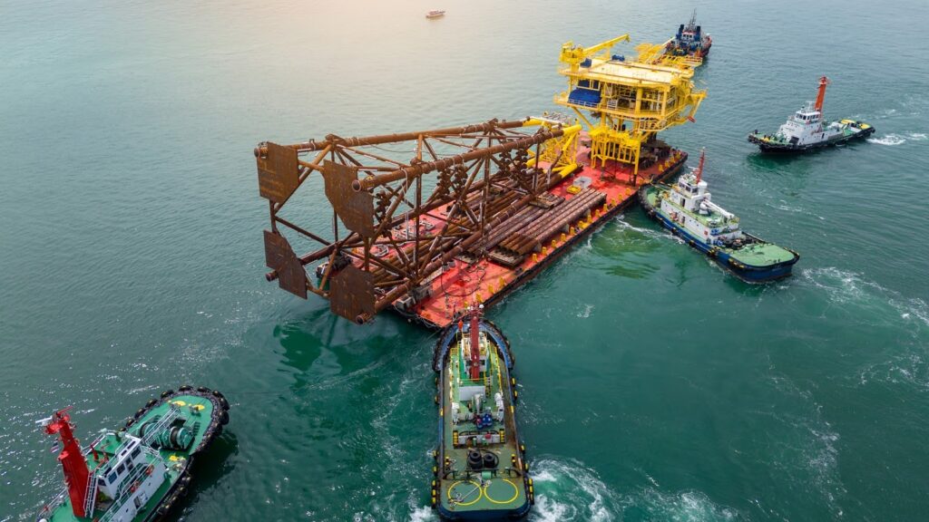 Four tugboats escort an oversized load—a massive offshore oil platform—on a barge along its purposed route that has been examined during a route survey. The yellow and brown structure dominates the deck, gliding over calm water under a slightly overcast sky.