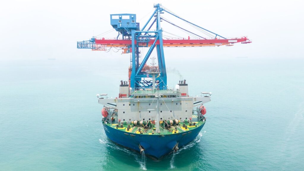 A large cargo ship with cranes docked on its deck sails along its purposed route, carrying an oversized load through calm turquoise waters under a hazy sky.
