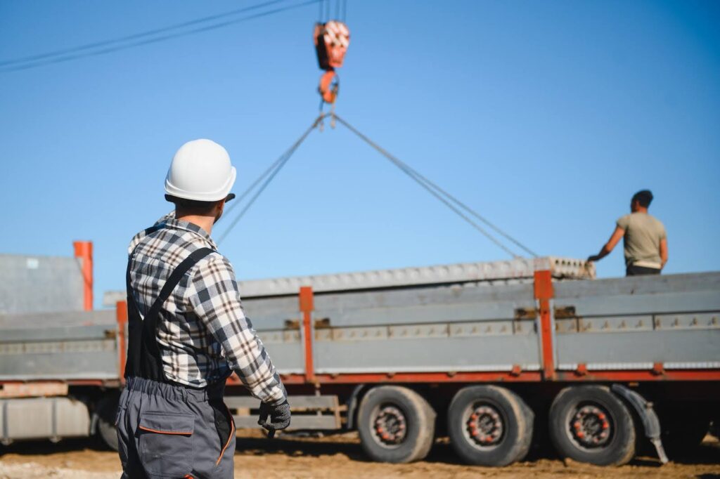 A construction worker in a white hard hat oversees a crane lifting concrete beams from a flatbed truck, while another worker stands on the truck, both under a clear blue sky.