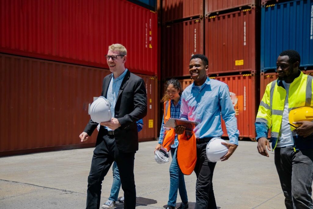 Four people, wearing business and safety attire, walk together smiling in front of large stacked shipping containers at a port. Two hold white hard hats, and one wears a high-visibility jacket.