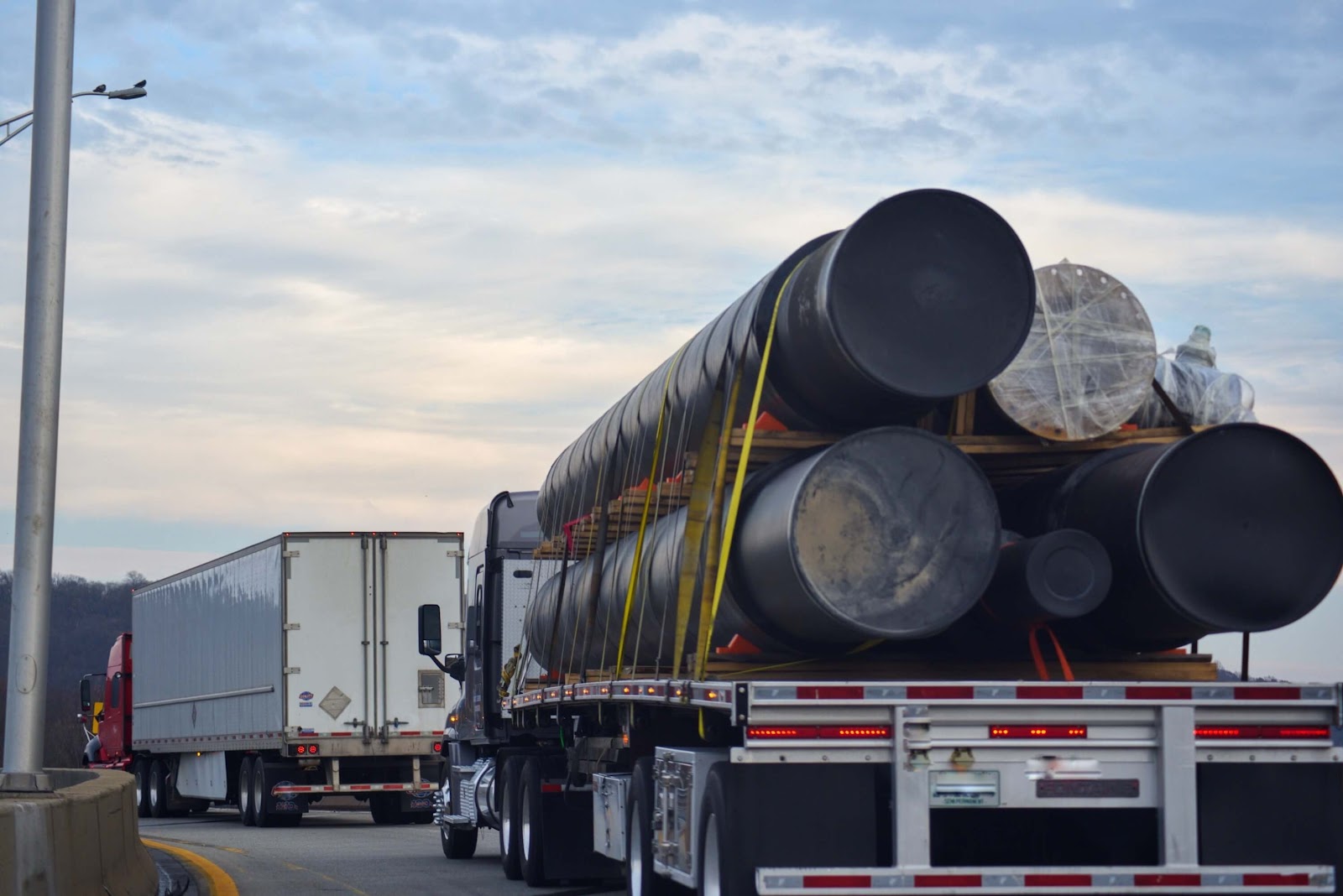 A flatbed truck hauling heavy haul loads of large cylindrical pipes, secured with straps, follows a white box truck on a highway under a cloudy sky. Trees are visible in the background.