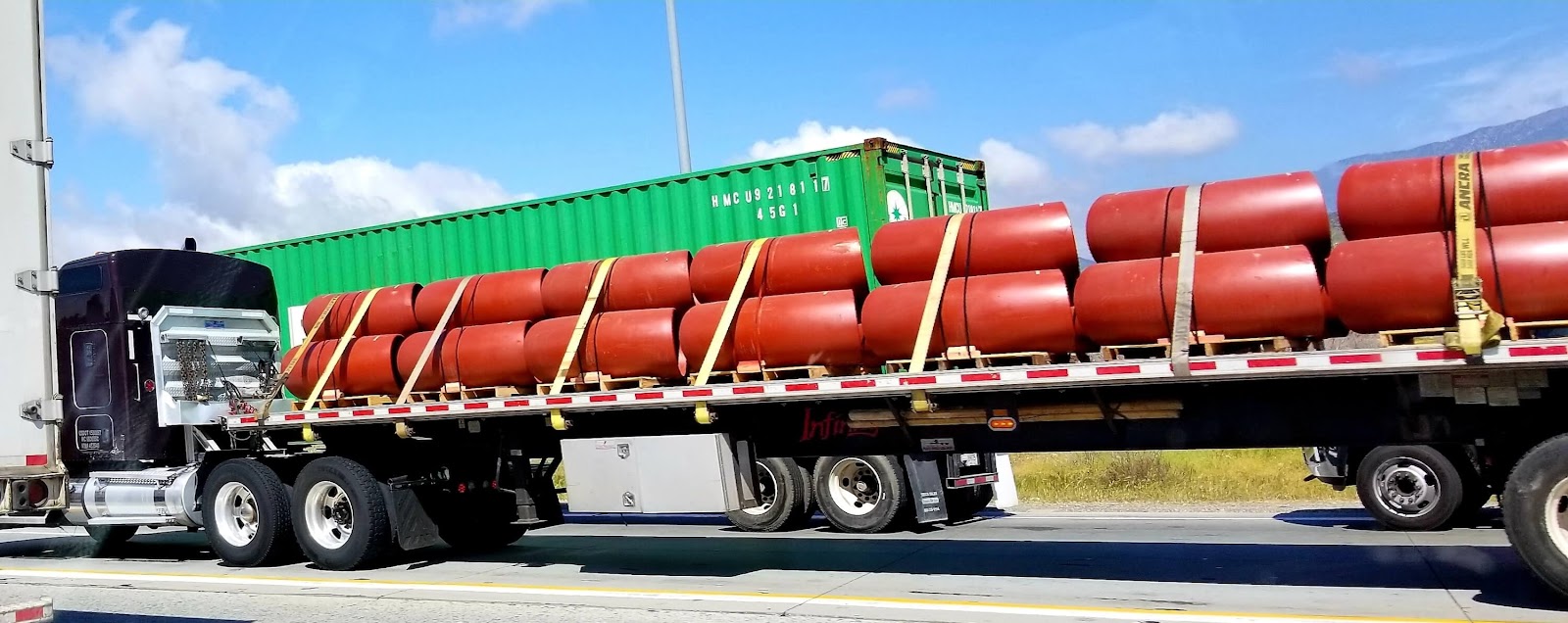 A flatbed semi-truck transports heavy haul loads—large, cylindrical red tanks secured with straps. In the background, a green shipping container sits on another trailer, and another truck is partially visible along the busy highway.