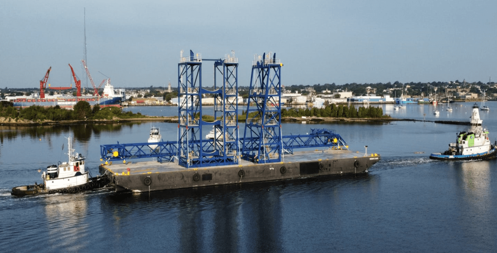 A large blue industrial structure, a typical example of heavy haul loads, is transported on a barge across calm water, assisted by two tugboats. Industrial cranes and buildings are visible in the background along the shoreline.
