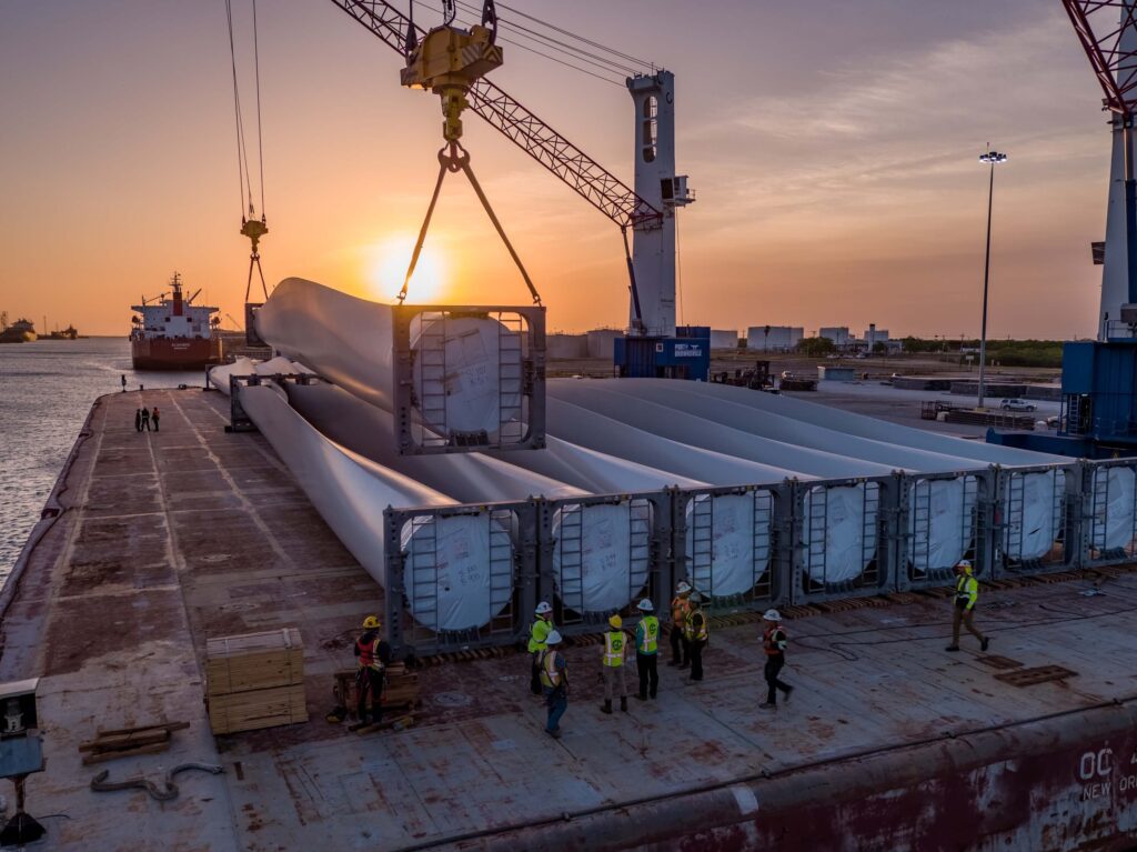 A crane lifts a long wind turbine blade onto a cargo ship at sunset. Workers in safety gear stand on deck as several heavy haul loads are already secured. Industrial port facilities are visible in the background. This is an example of how heavy haul freight rates work.