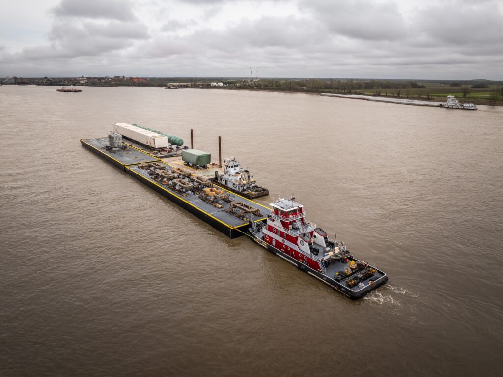 A red and white tugboat pushes a long barge carrying large industrial equipment and containers, illustrating oversized cargo shipping overseas along a wide, brown river under a cloudy sky. The shoreline and distant structures are visible in the background.