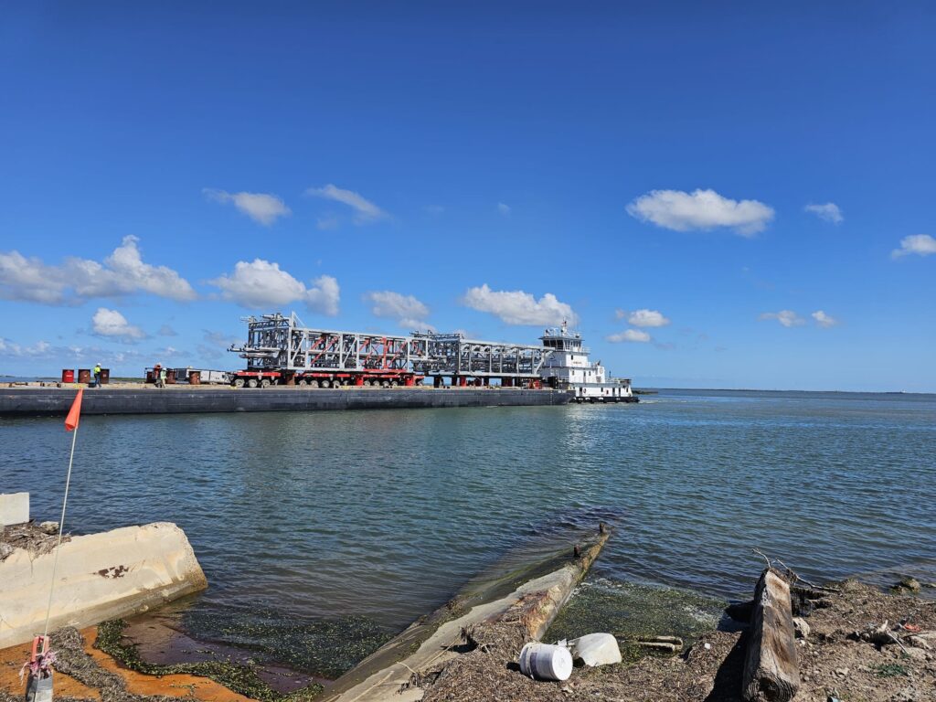 A barge carrying shipping machinery moves along a wide river under a blue sky with scattered clouds. The foreground shows debris and shoreline, while calm water reflects the sky's hues. It's an example of oversized cargo shipping overseas.