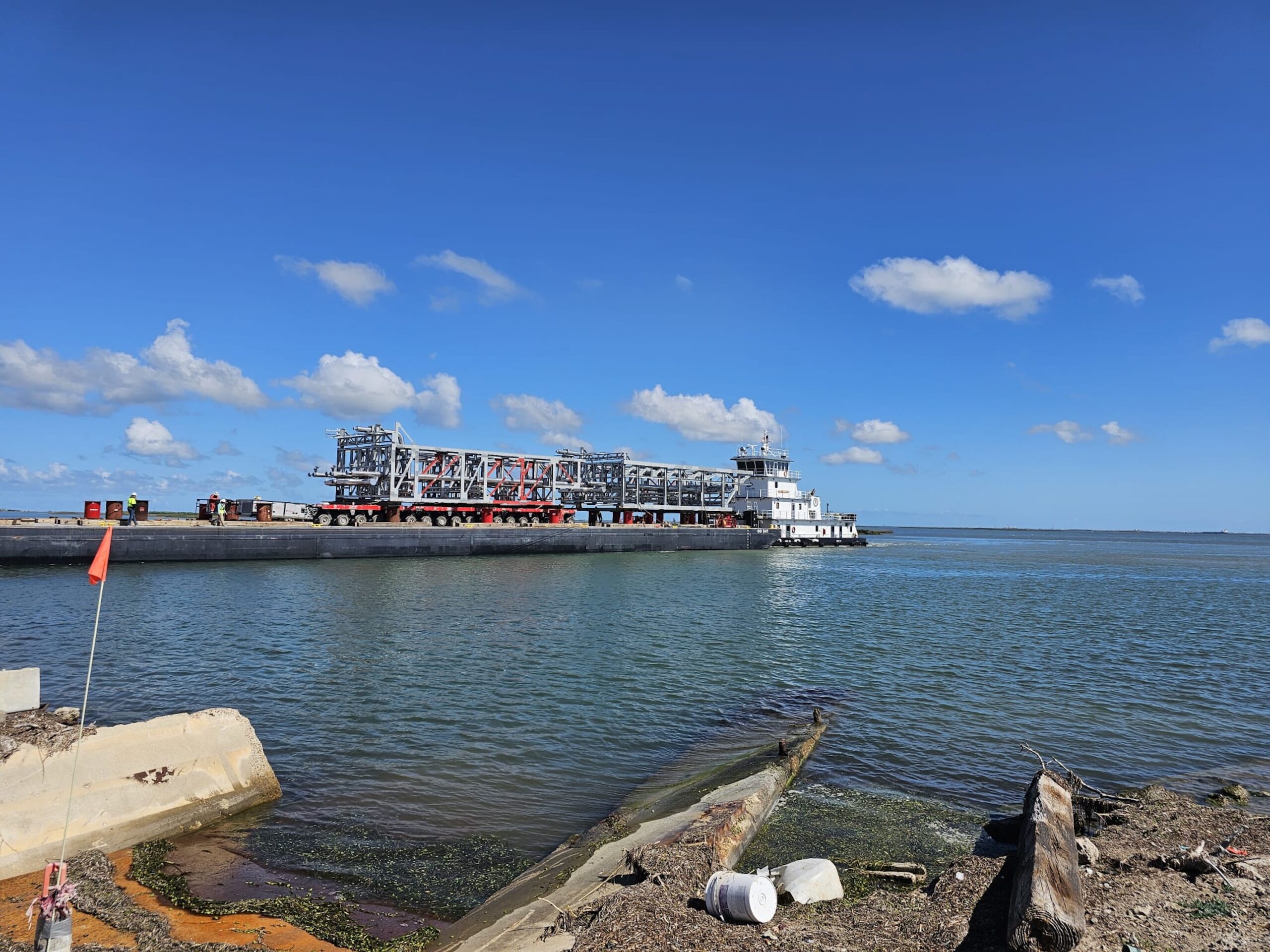 A barge carrying shipping machinery moves along a wide river under a blue sky with scattered clouds. The foreground shows debris and shoreline, while calm water reflects the sky's hues. It's an example of oversized cargo shipping overseas.