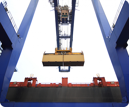 A large crane lifts a yellow shipping container above a cargo ship at a port, framed by blue metal structures against a bright sky.