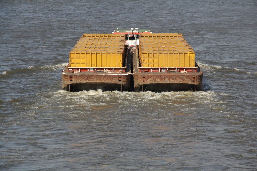 A barge carrying multiple large yellow containers—likely for shipping oversized cargo—moves through brownish river water, leaving a foamy wake behind it.