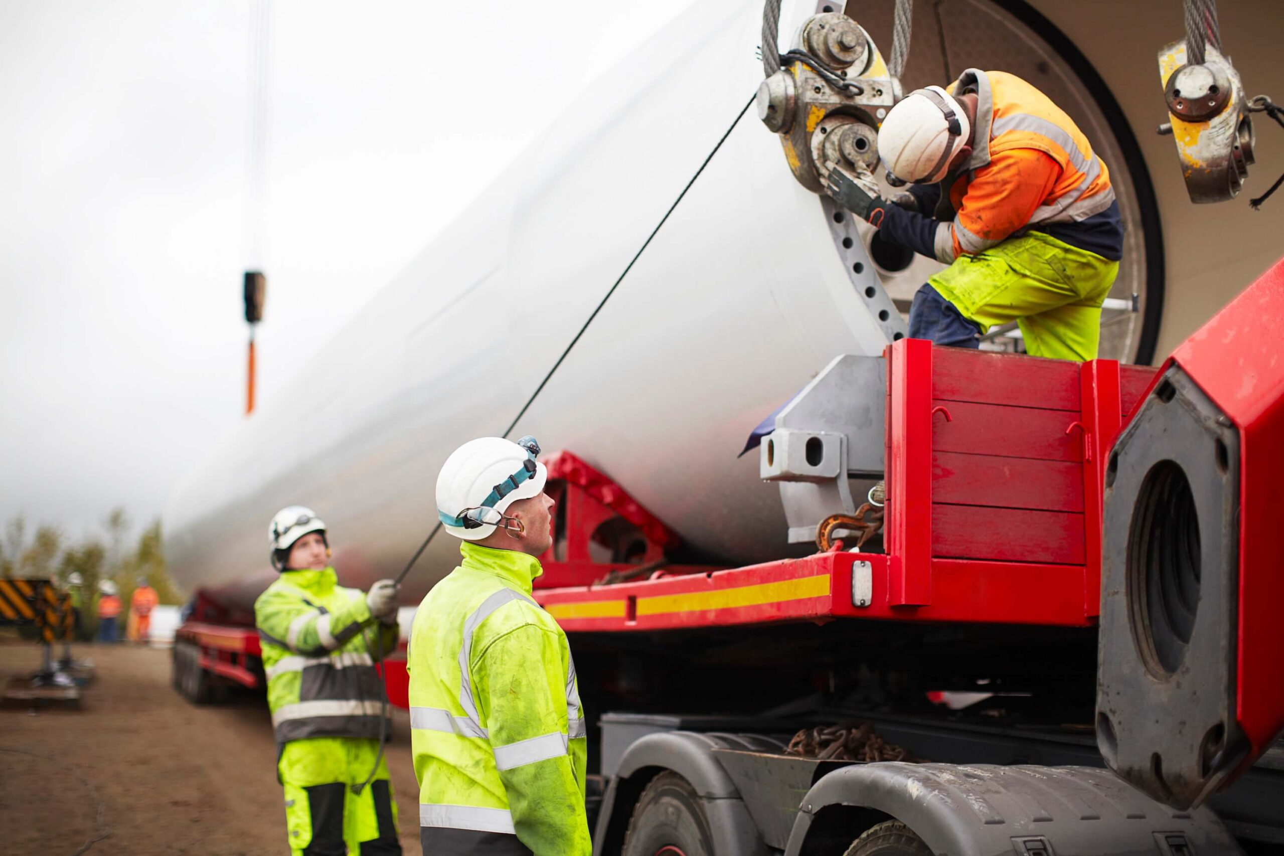 Three construction workers in safety gear load a large wind turbine component onto a red truck trailer using cranes and cables at an outdoor work site, preparing for oversized load route planning.