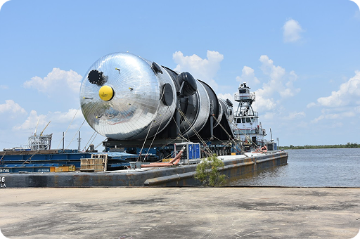 A large, cylindrical metal industrial vessel is loaded onto a barge at a dock, with water and a tugboat in the background under a partly cloudy blue sky.