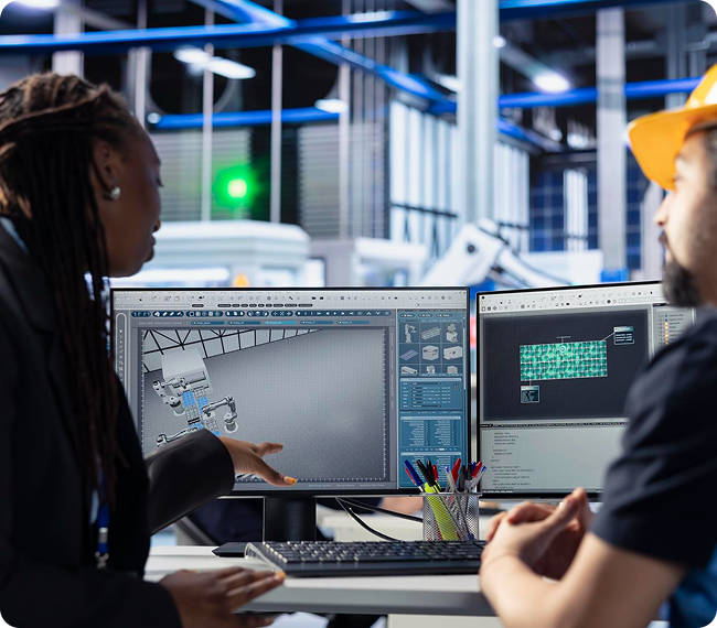Two people discuss 3D computer-aided design (CAD) models displayed on dual monitors in a modern industrial or engineering office setting. One points to the screen while the other listens attentively.