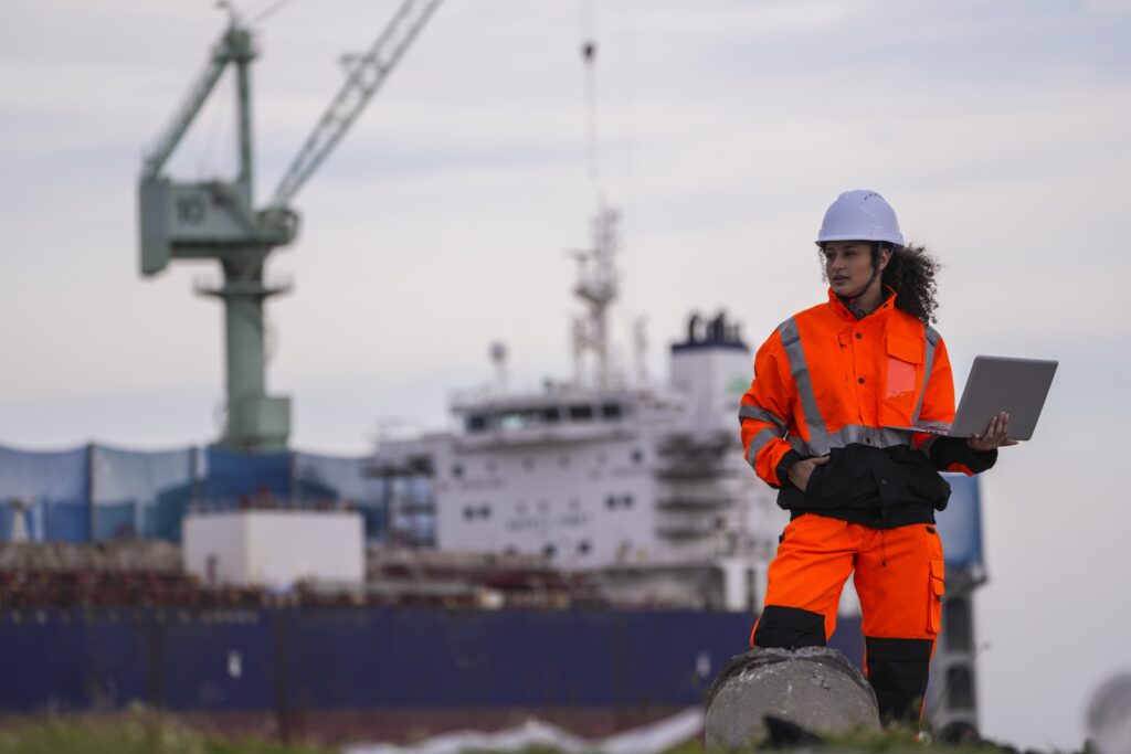 A person in a bright orange safety suit and white helmet stands outdoors holding a laptop, with a large cargo ship and cranes used for shipping oversized cargo visible in the background at a port or shipyard.