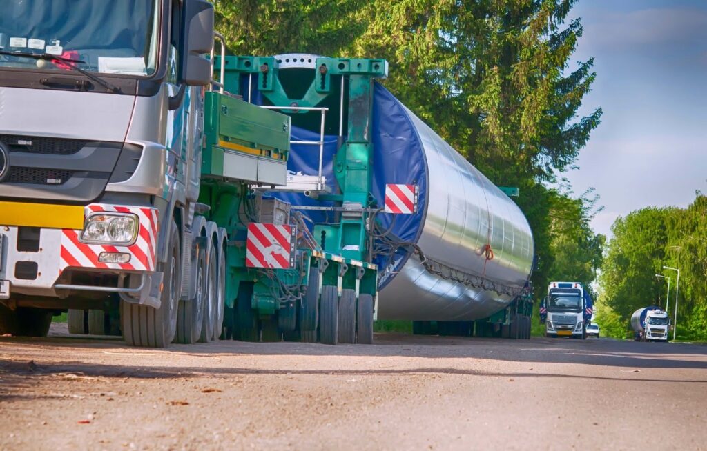 A large truck handles oversize loads, transporting a long, cylindrical wind turbine blade on a rural road, with two escort vehicles following behind. Trees line both sides of the road under a partly cloudy sky.