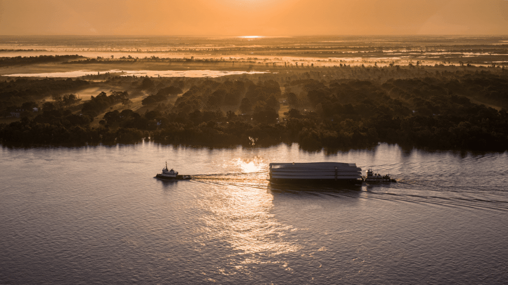 A tugboat pulls a large barge with an oversized freight shipping across a wide river at sunset, golden light reflecting on the water and a forested shoreline in the background.