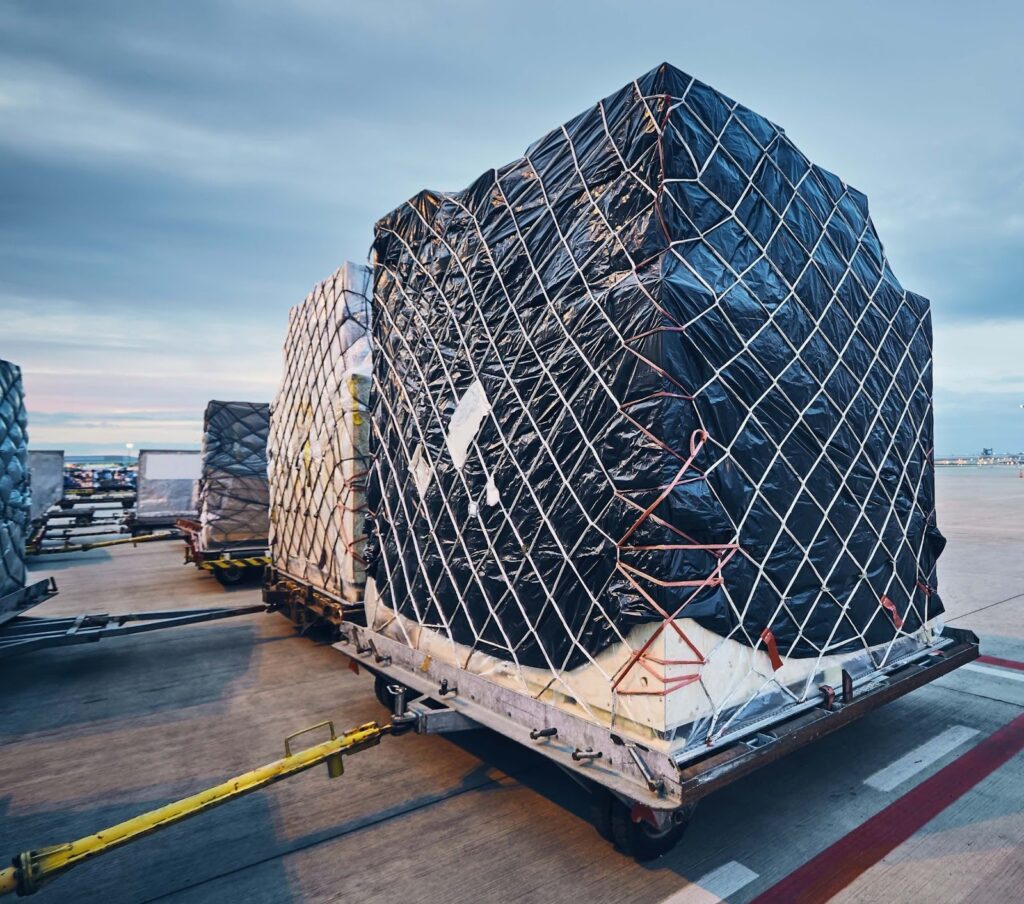 Large cargo wrapped in black plastic and secured with a net sits on a pallet trailer on an airport tarmac, ready for air transport. This oversized freight shipping operation highlights the challenges of handling oversize loads under strict weight limits.