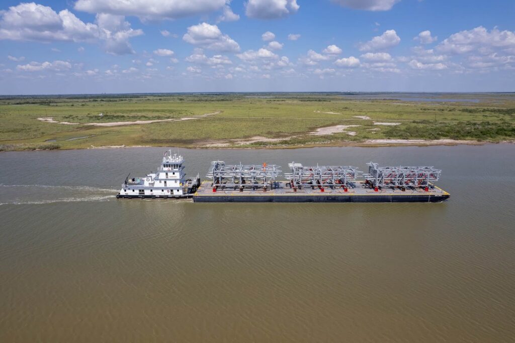 A barge carrying large metal structures as part of oversize load route planning is being pushed by a towboat along a wide river, with grassy land and a partly cloudy sky in the background.