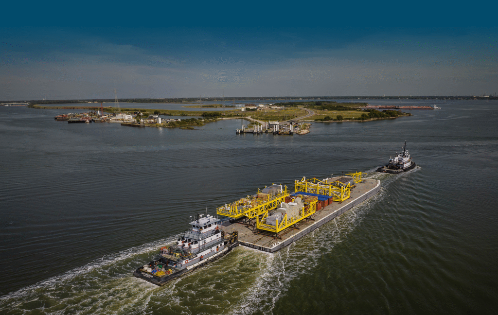 Aerial view of two tugboats towing a large barge loaded with industrial yellow equipment—possibly related to vessel repair duty exemptions—across a wide body of water, with a green island and distant shoreline in the background.