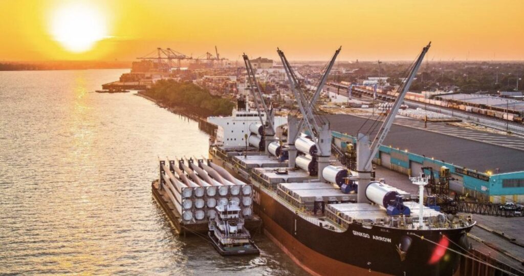 A large cargo ship loaded with industrial equipment is docked at a port during sunset, with cranes and storage buildings along the waterfront. The vibrant sky and water set the scene for secure project cargo insurance coverage.