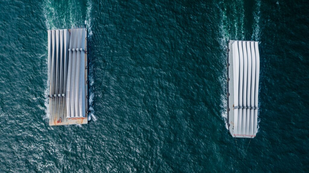 Aerial view of two large cargo ships engaged in project cargo transportation, carrying long, white structures as they travel parallel across deep blue-green water, leaving visible wakes behind them.