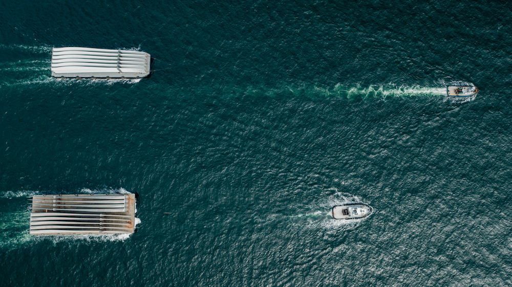 Aerial view of two large rectangular barges transporting project cargo and two smaller boats moving across deep blue water, leaving white wakes behind them. The vessels are parallel, spaced apart, and traveling in the same direction.