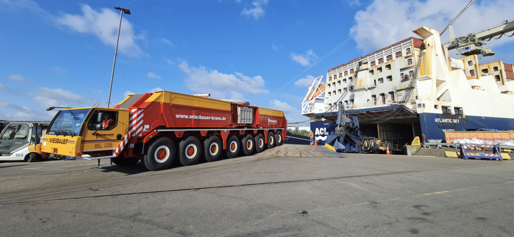 A large red and yellow multi-axle heavy transport vehicle prepares to board a cargo ship named "Atlantic Sky" at a port under a blue sky with scattered clouds.