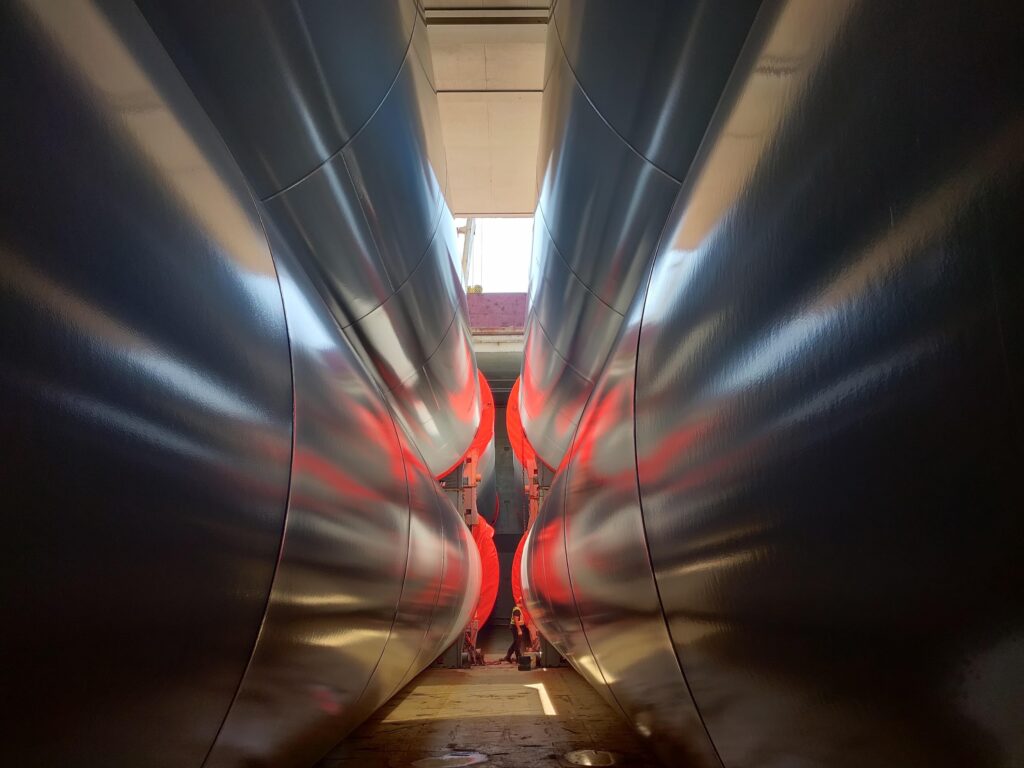 View between two large, curved, reflective metal surfaces with red light reflections, converging towards an open ceiling with daylight, and several small figures standing at the far end.