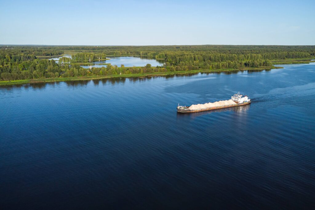 A cargo ship carrying goods, protected by cargo insurance, travels along a wide, calm river, with lush green trees and small lakes visible on the distant shoreline under a clear blue sky.