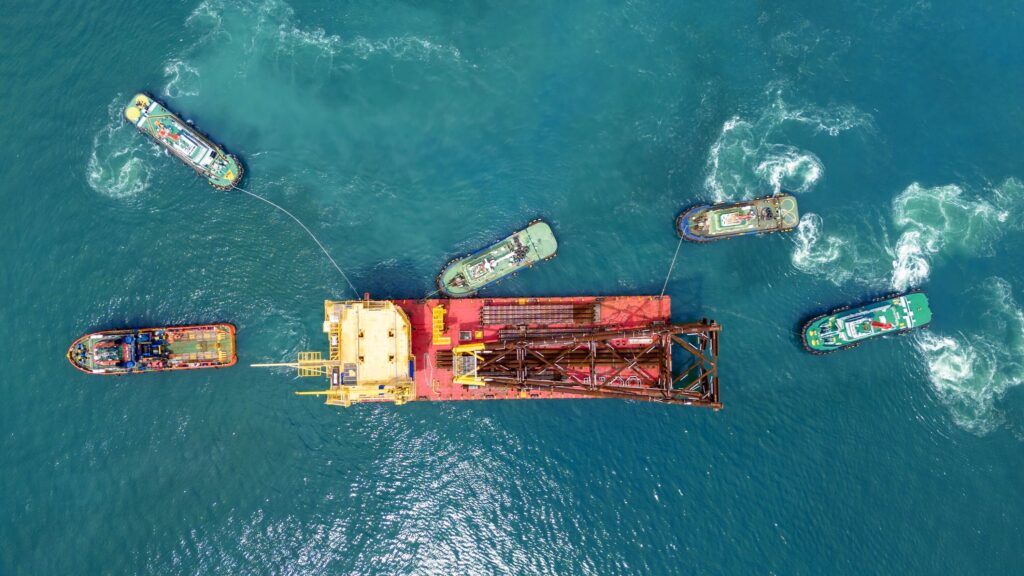 Aerial view of five tugboats maneuvering and towing a large red and yellow industrial barge in blue ocean waters, creating swirling patterns—an impressive display often safeguarded by marine cargo insurance.