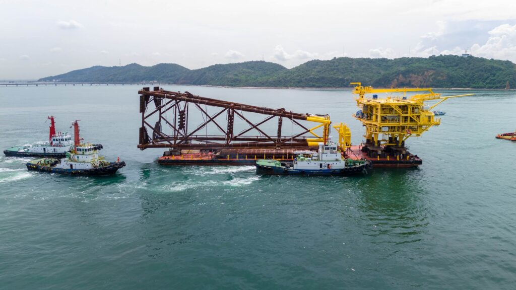 An offshore construction platform and a large metal structure are being transported on the water by several tugboats, with hilly land visible in the background under a cloudy sky—ideal scenarios highlighting the importance of marine cargo insurance.